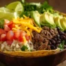 Taco bowl recipe featuring seasoned beef, rice, black beans, lettuce, and avocado on a rustic wooden table.